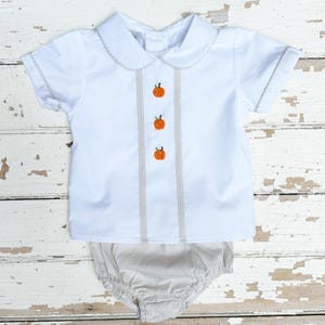 May include: White short-sleeved shirt and matching diaper cover set. The shirt features a Peter Pan collar, gray trim, and three embroidered orange pumpkins down the front. The diaper cover is a light brown and white gingham pattern.