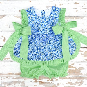 May include: A baby outfit featuring a blue floral print top with green gingham trim and bows. The outfit includes matching bloomers. The garment is designed with ruffles and a playful aesthetic, perfect for a summer day.