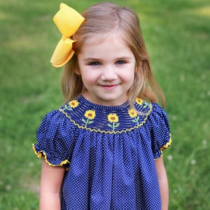 May include: A young girl wearing a navy blue polka dot dress with yellow sunflower embroidery and trim. She has a large yellow bow in her hair. The dress has short sleeves.