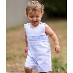 May include: A white romper with blue embroidered details. The sleeveless romper features blue button closures on the shoulders and a decorative embroidered band across the chest. The garment is designed for a baby or toddler.