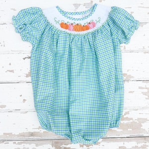 May include: A baby's romper in a blue and green gingham pattern. The neckline features an embroidered design of orange pumpkins. The short-sleeved romper has a gathered bodice and a bubble silhouette, perfect for fall.