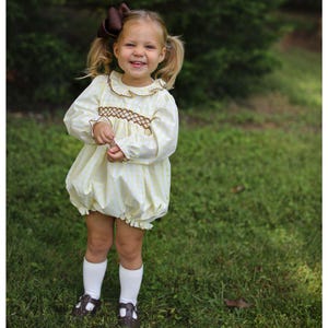 May include: A young child wearing a yellow and white striped romper with a brown bow in their hair. The romper has long sleeves, a ruffled collar, and brown embroidery. The child is also wearing white socks and brown shoes.