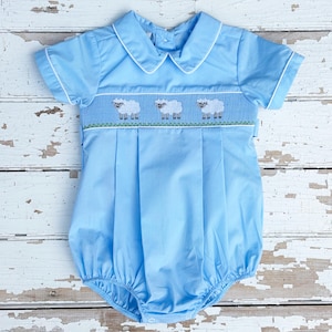 May include: Light blue baby romper with white trim and a Peter Pan collar. The romper features a smocked panel with three embroidered sheep above a green grass design. The short-sleeved romper has a pleated front and a bubble bottom.