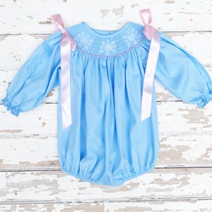 May include: A light blue baby romper with long sleeves and a round neckline. The neckline features white snowflake embroidery with pink accents. Pink satin ribbons are tied on the shoulders. The romper is made of a soft, lightweight fabric.