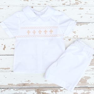 May include: A white two-piece outfit for a child. The short-sleeved shirt has a rounded collar and a decorative band with embroidered crosses. The matching shorts are white and have an elastic waistband. The outfit is made of a lightweight fabric.