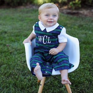 May include: A young child wearing a blue, green, and white plaid romper with the initials "WCL" embroidered on the front. The romper has a white collar and short sleeves. The child is sitting in a white chair on a grassy surface.
