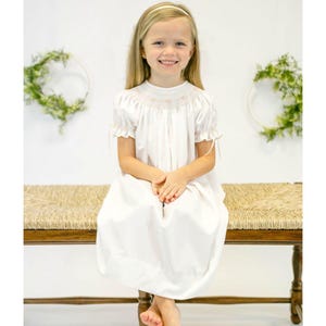 May include: A white dress with short sleeves and a smocked bodice. The dress has a round neckline and a full skirt. The sleeves are ruffled at the cuffs. The dress is worn by a young person with blonde hair and a headband, sitting on a wooden bench.