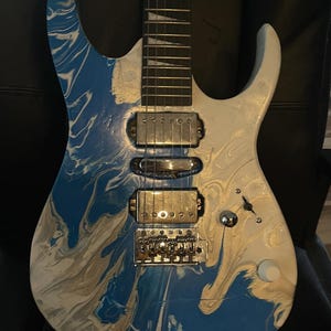 Puede incluir: Guitarra el&eacute;ctrica con acabado veteado azul, blanco y plateado. La guitarra tiene un m&aacute;stil blanco con trastes plateados y un puente y pastillas plateadas. El cuerpo presenta un patr&oacute;n arremolinado de azul, blanco y plateado. Es un instrumento de seis cuerdas.