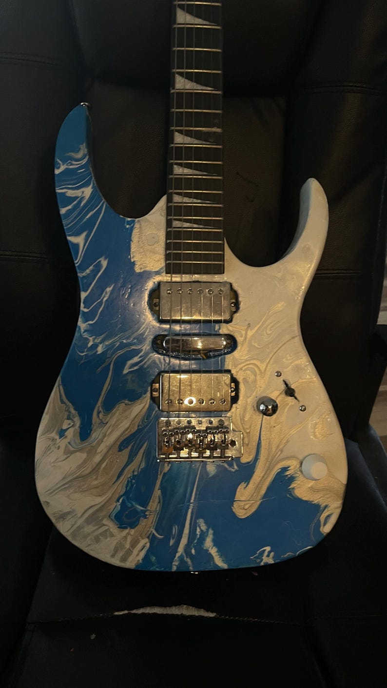Puede incluir: Guitarra el&eacute;ctrica con acabado veteado azul, blanco y plateado. La guitarra tiene un m&aacute;stil blanco con trastes plateados y un puente y pastillas plateadas. El cuerpo presenta un patr&oacute;n arremolinado de azul, blanco y plateado. Es un instrumento de seis cuerdas.