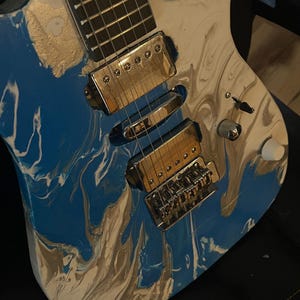Puede incluir: Guitarra el&eacute;ctrica con acabado veteado en azul, blanco y dorado. La guitarra presenta herrajes cromados, incluyendo pastillas, puente y clavijas de afinaci&oacute;n. El diapas&oacute;n es oscuro con trastes plateados. El cuerpo tiene un dise&ntilde;o de pintura &uacute;nico y arremolinado.