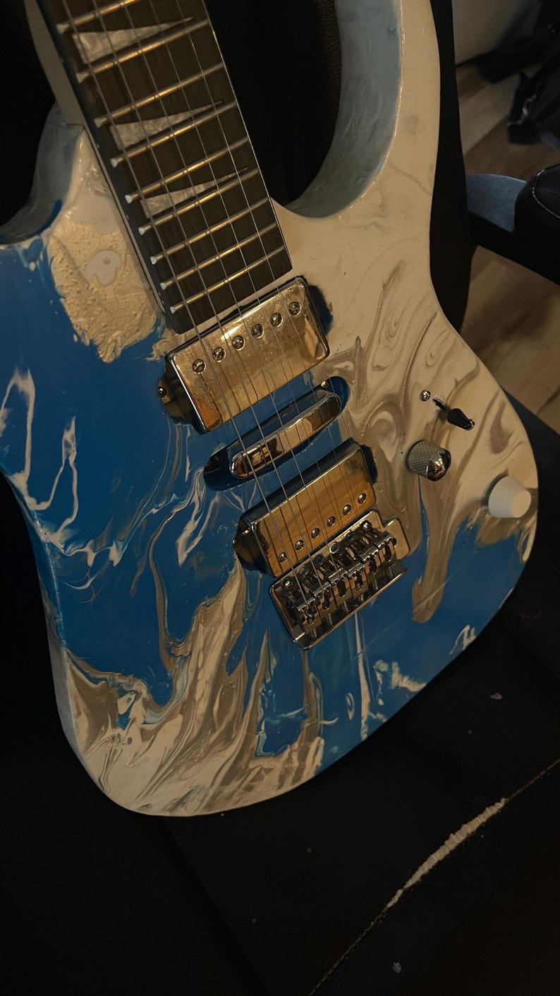 Puede incluir: Guitarra el&eacute;ctrica con acabado veteado en azul, blanco y dorado. La guitarra presenta herrajes cromados, incluyendo pastillas, puente y clavijas de afinaci&oacute;n. El diapas&oacute;n es oscuro con trastes plateados. El cuerpo tiene un dise&ntilde;o de pintura &uacute;nico y arremolinado.
