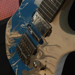 Puede incluir: Guitarra el&eacute;ctrica con acabado veteado azul, blanco y gris. La guitarra presenta herrajes cromados, incluido el puente, las pastillas y los clavijeros. El diapas&oacute;n es oscuro con incrustaciones blancas. Se ve un bot&oacute;n de volumen blanco.