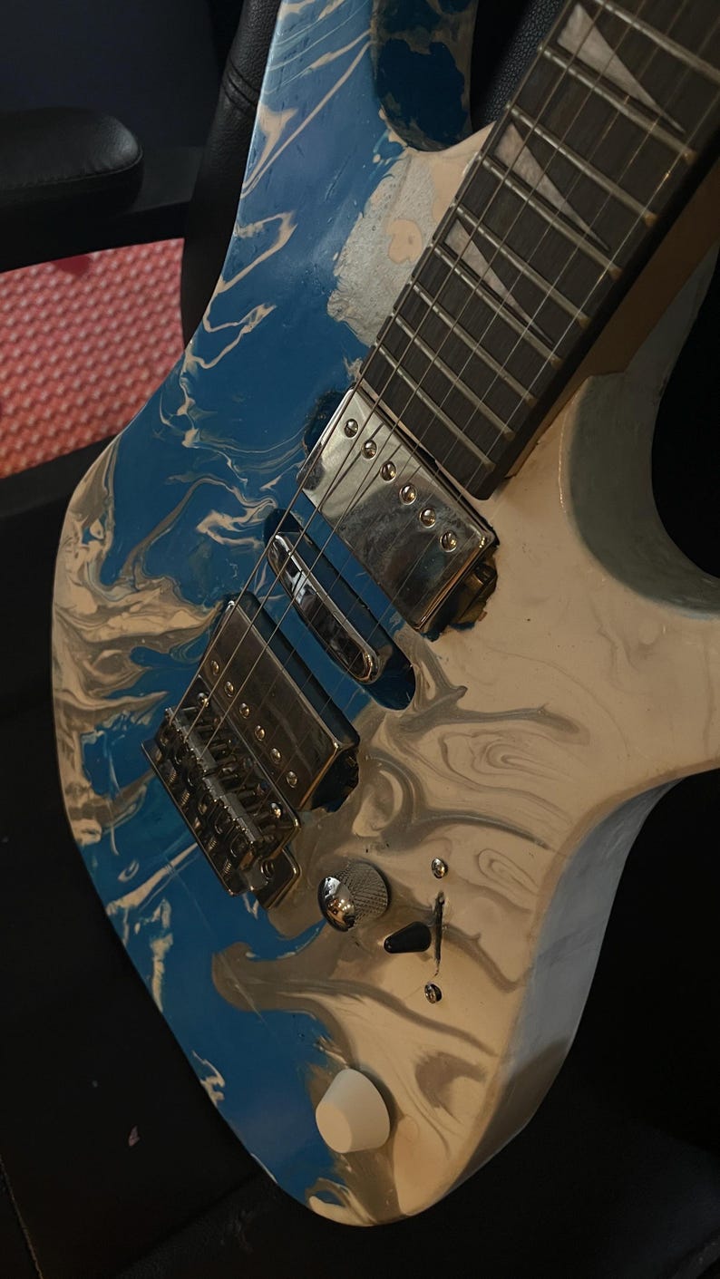 Puede incluir: Guitarra el&eacute;ctrica con acabado veteado azul, blanco y gris. La guitarra presenta herrajes cromados, incluido el puente, las pastillas y los clavijeros. El diapas&oacute;n es oscuro con incrustaciones blancas. Se ve un bot&oacute;n de volumen blanco.