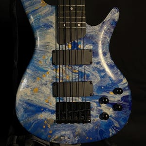 Könnte beinhalten: Ein sechssaitiger Bassgitarre mit einem wirbelnden Muster in Blau, Weiß und Gold. Das Instrument hat schwarze Hardware, einschließlich der Tonabnehmer, der Brücke und der Stimmwirbel. Ein schwarzer Gurt ist angebracht. Der Bass steht auf einem Ständer vor schwarzem Hintergrund.