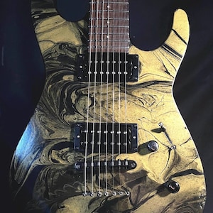 Puede incluir: Guitarra eléctrica con un acabado en espiral dorado y negro. La guitarra tiene pastillas, perillas y un diapasón negros. El clavijero tiene el texto "E-II 17". El instrumento se muestra sobre un fondo negro.