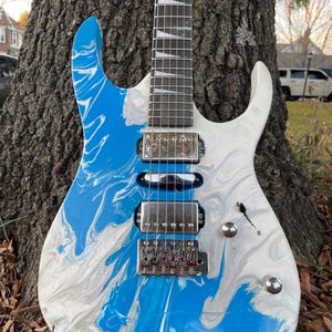 Puede incluir: Guitarra el&eacute;ctrica con acabado veteado azul y blanco. La guitarra tiene un puente, pastillas y clavijas plateadas. El diapas&oacute;n es oscuro con incrustaciones blancas. El cuerpo est&aacute; apoyado contra un tronco de &aacute;rbol.