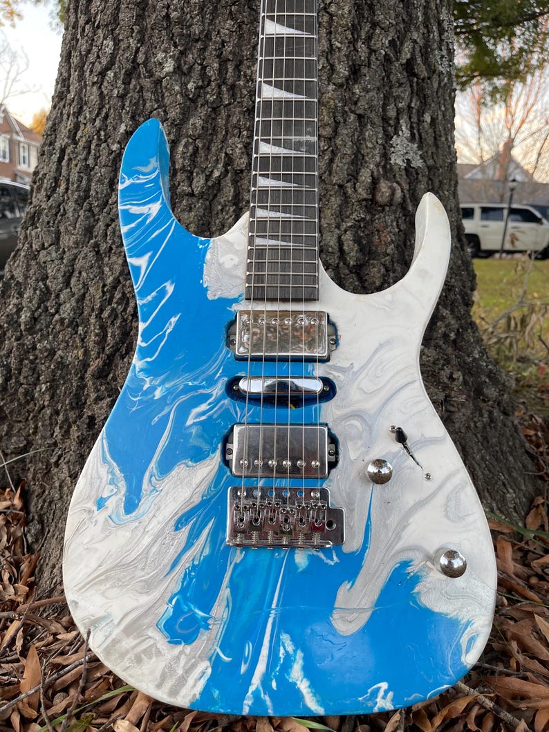 Puede incluir: Guitarra el&eacute;ctrica con acabado veteado azul y blanco. La guitarra tiene un puente, pastillas y clavijas plateadas. El diapas&oacute;n es oscuro con incrustaciones blancas. El cuerpo est&aacute; apoyado contra un tronco de &aacute;rbol.