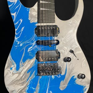 Puede incluir: Guitarra el&eacute;ctrica con un patr&oacute;n de remolino azul, blanco y plateado en el cuerpo. La guitarra tiene un m&aacute;stil plateado, pastillas negras y herrajes plateados. El clavijero es blanco con clavijas negras.