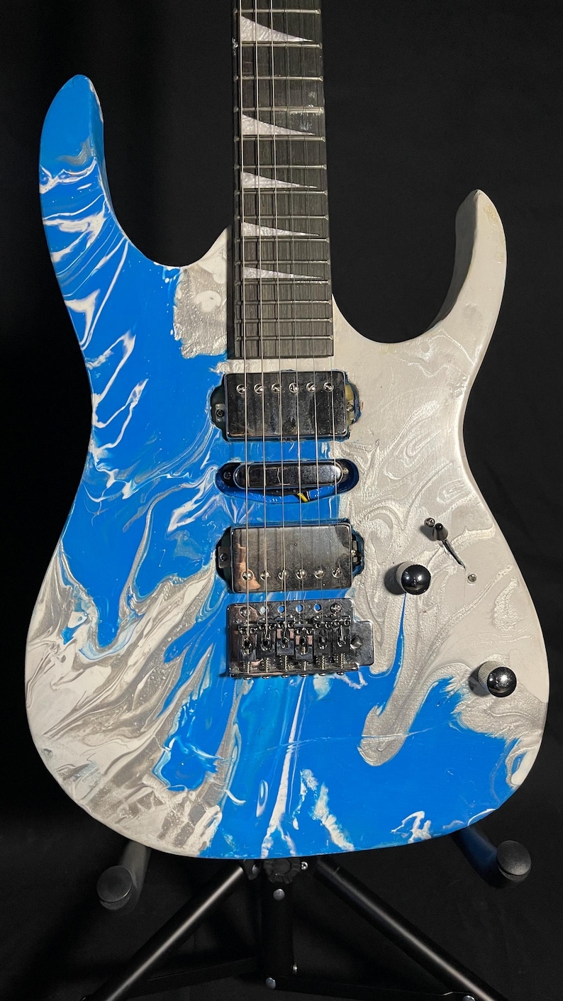 Puede incluir: Guitarra el&eacute;ctrica con un patr&oacute;n de remolino azul, blanco y plateado en el cuerpo. La guitarra tiene un m&aacute;stil plateado, pastillas negras y herrajes plateados. El clavijero es blanco con clavijas negras.