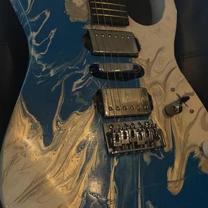 Puede incluir: Guitarra el&eacute;ctrica con un acabado en espiral azul, blanco y dorado. La guitarra tiene herrajes cromados, incluyendo pastillas, un puente y clavijas de afinaci&oacute;n. El cuerpo tiene un dise&ntilde;o de doble corte.