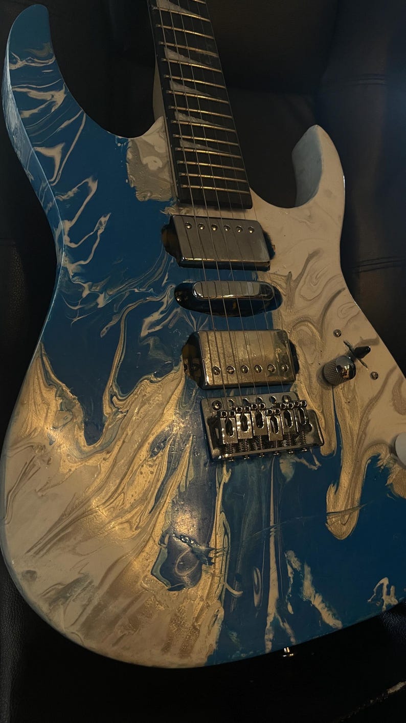 Puede incluir: Guitarra el&eacute;ctrica con un acabado en espiral azul, blanco y dorado. La guitarra tiene herrajes cromados, incluyendo pastillas, un puente y clavijas de afinaci&oacute;n. El cuerpo tiene un dise&ntilde;o de doble corte.