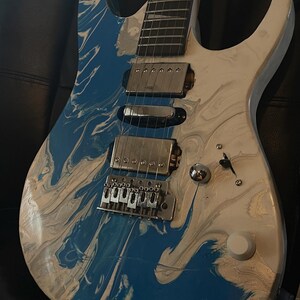 Puede incluir: Guitarra el&eacute;ctrica con acabado veteado azul y blanco. La guitarra tiene un puente, pastillas y clavijas plateadas. El cuerpo es una mezcla de remolinos azules y blancos, y el m&aacute;stil es de color madera oscura.