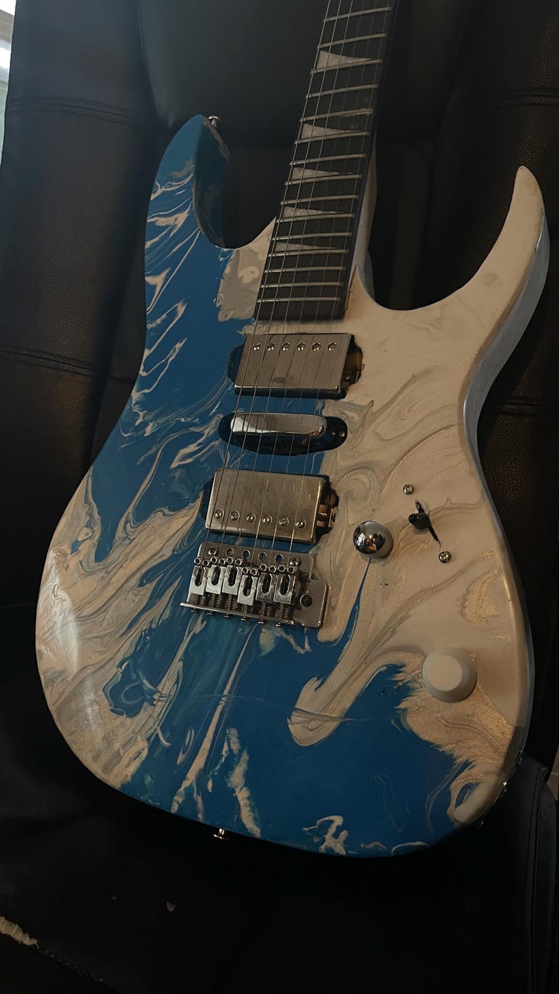 Puede incluir: Guitarra el&eacute;ctrica con acabado veteado azul y blanco. La guitarra tiene un puente, pastillas y clavijas plateadas. El cuerpo es una mezcla de remolinos azules y blancos, y el m&aacute;stil es de color madera oscura.