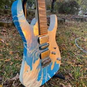 Puede incluir: Una guitarra el&eacute;ctrica con un acabado veteado azul, blanco y dorado &uacute;nico. La guitarra est&aacute; montada en un soporte negro, mostrando el dise&ntilde;o de su cuerpo. El diapas&oacute;n y los herrajes son visibles, destacando los detalles del instrumento.