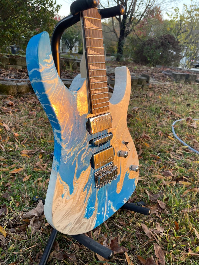 Puede incluir: Una guitarra el&eacute;ctrica con un acabado veteado azul, blanco y dorado &uacute;nico. La guitarra est&aacute; montada en un soporte negro, mostrando el dise&ntilde;o de su cuerpo. El diapas&oacute;n y los herrajes son visibles, destacando los detalles del instrumento.