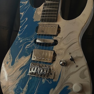 Puede incluir: Guitarra el&eacute;ctrica con acabado veteado azul y blanco. La guitarra tiene un puente, pastillas y clavijas plateadas. El cuerpo es una mezcla de remolinos azules y blancos, creando un efecto visual &uacute;nico. La guitarra est&aacute; sobre una superficie negra.