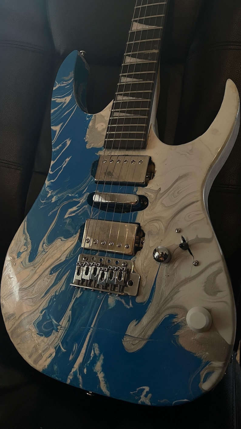 Puede incluir: Guitarra el&eacute;ctrica con acabado veteado azul y blanco. La guitarra tiene un puente, pastillas y clavijas plateadas. El cuerpo es una mezcla de remolinos azules y blancos, creando un efecto visual &uacute;nico. La guitarra est&aacute; sobre una superficie negra.