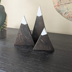 May include: Three wooden mountain sculptures with dark brown stained bodies and white painted peaks. The decorative tabletop items are arranged in a triangular formation, creating a minimalist, nature-inspired aesthetic.