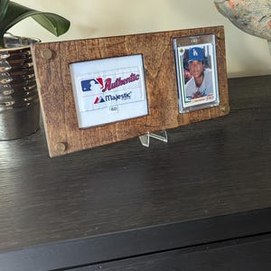 May include: A wooden baseball card display frame with two openings. One holds a patch with "Authentic Majestic" text, the other a baseball card. The frame is brown and has a metal stand.