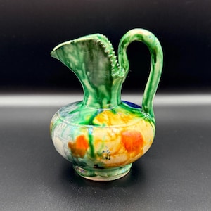 May include: A small, hand-painted ceramic pitcher with a rounded body and a curved handle. The pitcher features a vibrant color palette of green, yellow, orange, and blue. The spout has a decorative, textured edge. The pitcher is likely for decorative use.