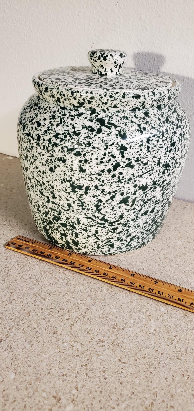 May include: A ceramic jar with a lid, featuring a white base and a speckled pattern of dark green. The jar is approximately 10 inches tall and 8 inches wide. A wooden ruler is placed next to the jar.