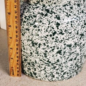 May include: A ceramic jar with a lid, featuring a white base and a speckled green pattern. The jar is approximately 9 inches tall, with a matching lid and a small knob. A wooden ruler is placed next to the jar.