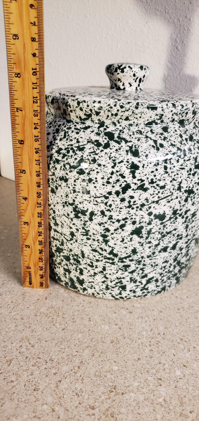 May include: A ceramic jar with a lid, featuring a white base and a speckled green pattern. The jar is approximately 9 inches tall, with a matching lid and a small knob. A wooden ruler is placed next to the jar.