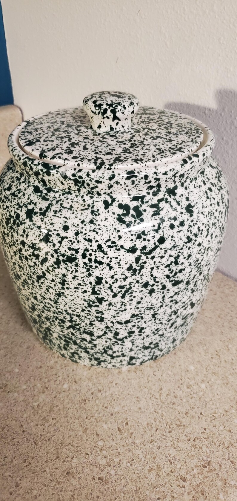 May include: A ceramic jar with a lid, featuring a speckled pattern of dark green spots on a white background. The jar has a rounded shape and a small knob on the lid. The jar is likely used for storage.