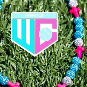 May include: A beaded necklace with blue and silver rhinestone beads, pink cross-shaped beads, and a Miami Ice logo. The necklace is set against a backdrop of green grass.