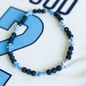 May include: A necklace featuring dark blue, sparkly beads and light blue cross-shaped beads. The necklace is on a white background with the word "WOOD" and the number "2" in blue.