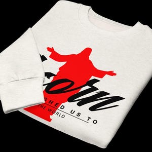 May include: A white long-sleeve sweatshirt featuring a red silhouette of a figure with outstretched arms. The black text reads "KORN" in a large script font, with "LAUGHED US TO THE WORLD" printed in smaller capital letters underneath.