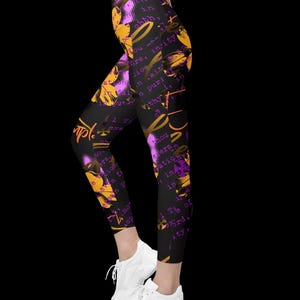 May include: Black leggings featuring a vibrant graphic print with orange and purple floral motifs, abstract shapes, and scattered text in various fonts. The design includes repeating words and phrases layered over the dark fabric, paired with white sneakers.