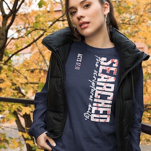May include: Navy blue long-sleeve shirt with the text "SEARCHED the scriptures daily" in a vertical design. The word "SEARCHED" is in a red and white marble design. The text "ACTS 17:11" is in white. The person is wearing a black vest.