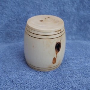 May include: A small, barrel-shaped wooden container with a lid. The light-coloured wood has dark brown bands and a knot detail on the side. The container is likely used for storage or decorative purposes.