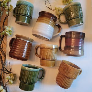 May include: A selection of ceramic mugs in various colours and designs. The mugs feature shades of green, brown, and tan, with textured and patterned surfaces. Some mugs have a stacked design, while others have a more traditional shape. The mugs are arranged on a white surface.