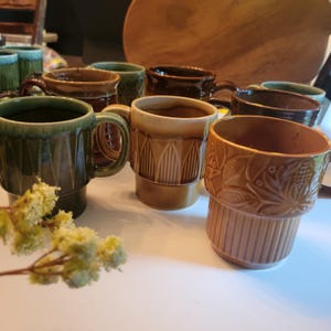 May include: A collection of ceramic mugs in various shades of brown and green. The mugs have unique textured designs, including vertical lines and floral patterns. Each mug has a handle and a cylindrical shape, perfect for enjoying hot beverages.