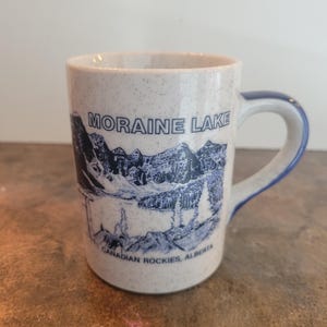 Peut inclure: Une tasse en céramique mouchetée avec une illustration bleue et blanche du lac Moraine dans les Rocheuses canadiennes. La tasse a une anse et un bord bleus, avec les mots "Moraine Lake" et "Canadian Rockies, Alberta" imprimés dessus.