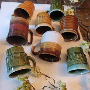 May include: A collection of ceramic mugs in various colors and designs. The mugs feature shades of brown, green, and white, with textured patterns and handles. Some mugs have a layered color scheme. The mugs are arranged on a white surface.