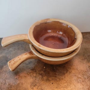 May include: Two stacked, brown ceramic bowls with handles. The bowls have a glossy interior and a lighter rim. The handles are a lighter colour with a speckled pattern. The bowls are likely for serving soup or other dishes.