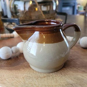 May include: A small, ceramic creamer with a brown rim and handle, a tan stripe, and a cream-coloured base. The creamer has a spout for easy pouring and a rounded body. This kitchenware item is suitable for serving cream or milk.
